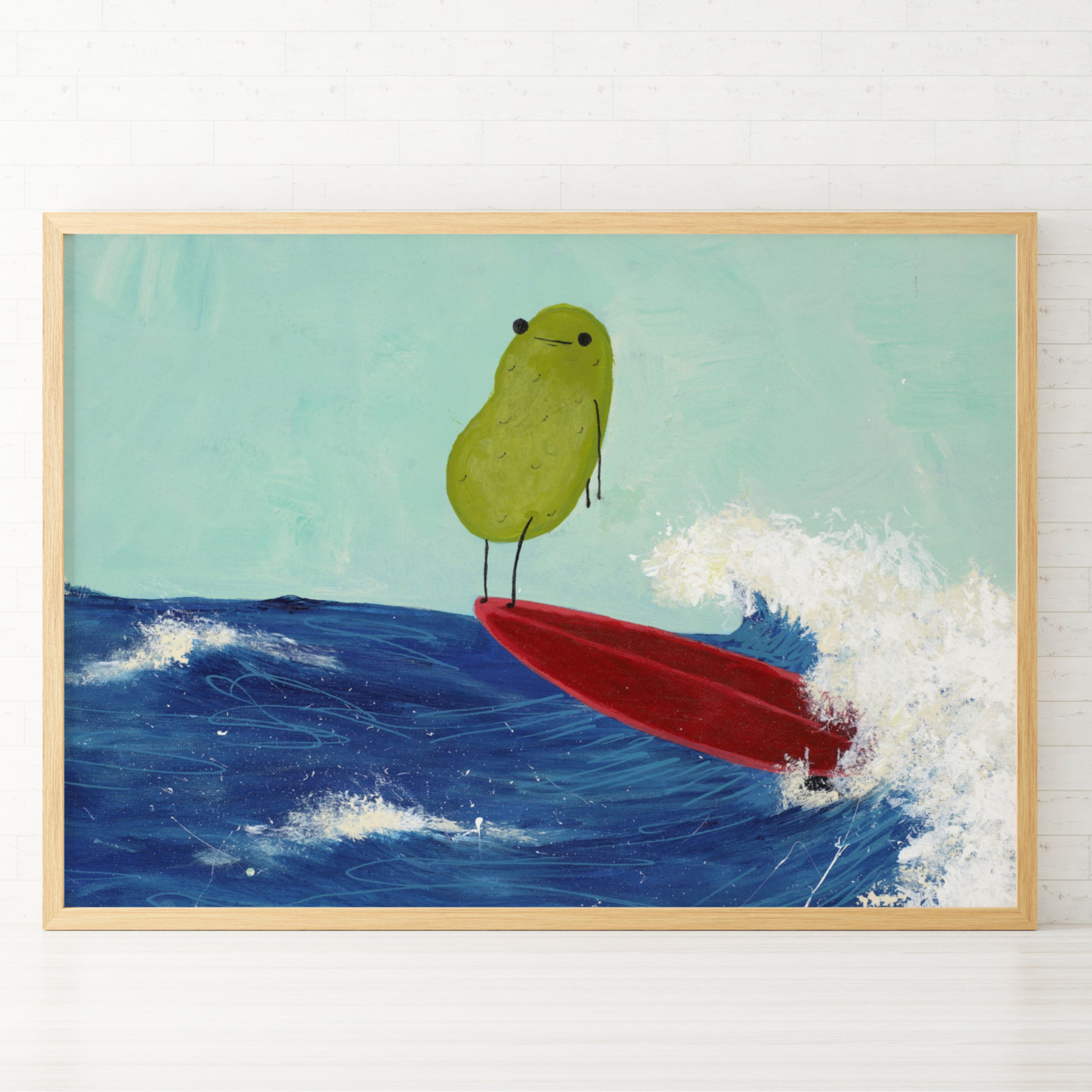 a pickle art print featuring a cute pickle riding a red surfboard on a wave is displayed in a wooden frame against a white tiled wall