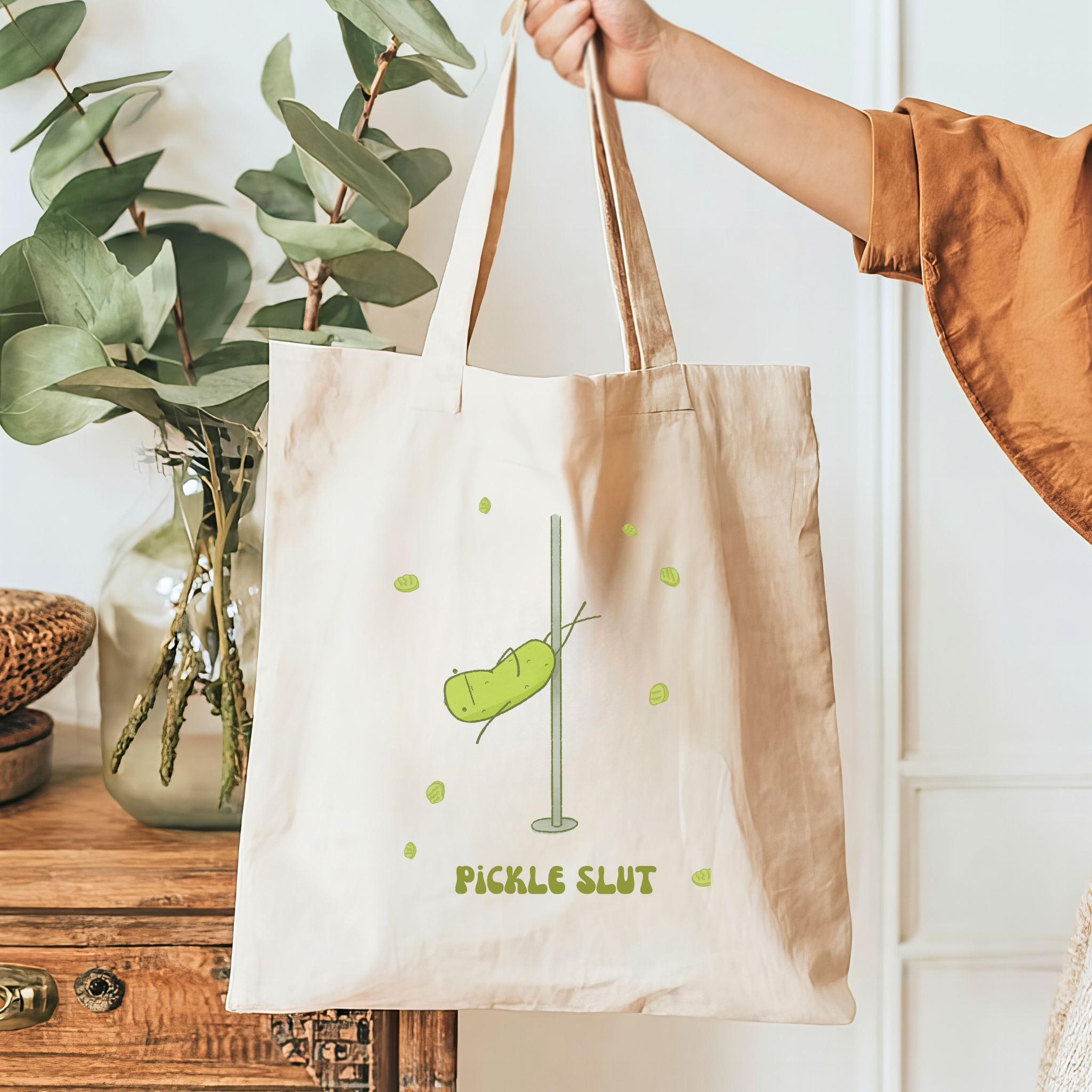 Tote bag with 'Pickle Slut' design held by a person, with a plant and wooden cabinet in the background.