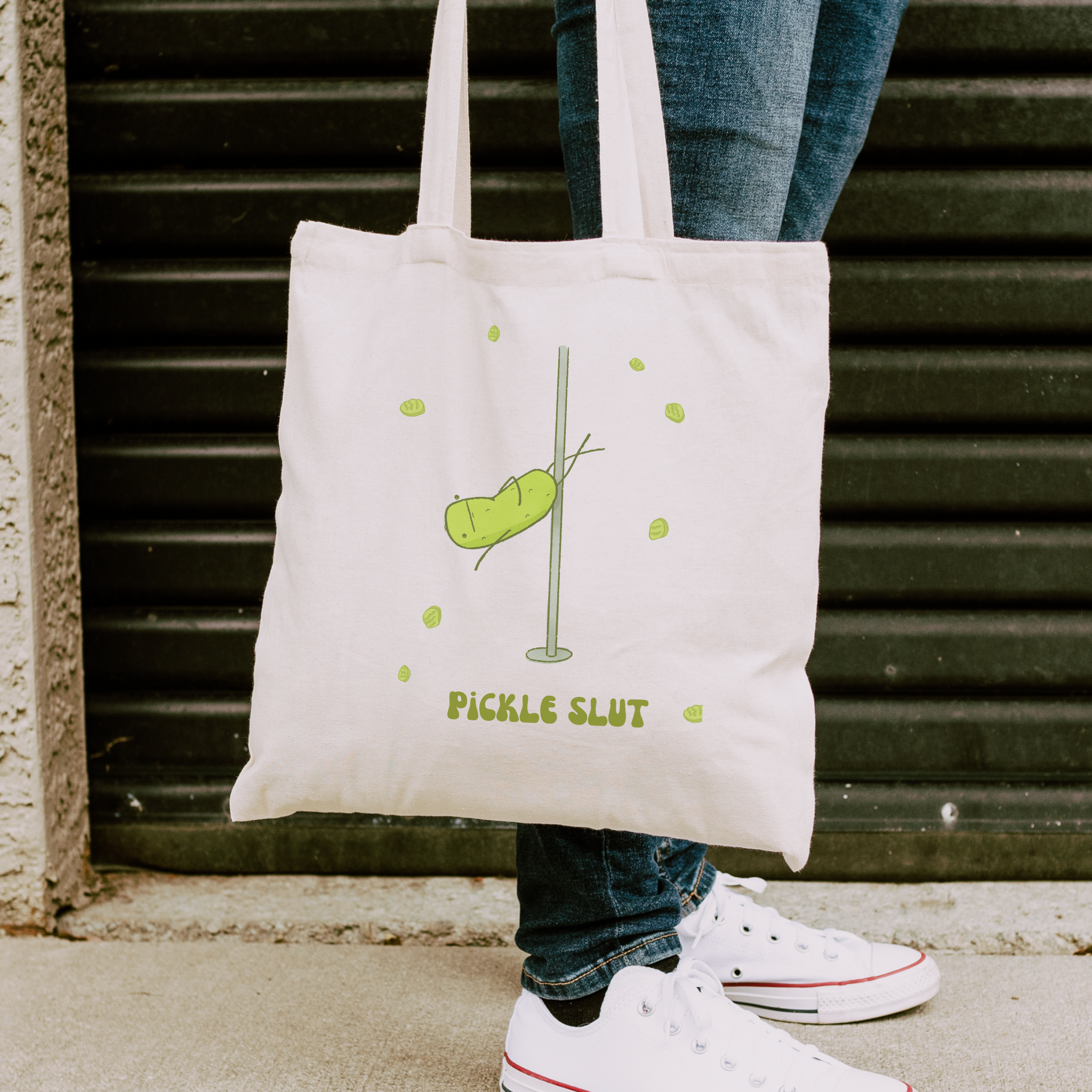 person holding a pickle slut tote bag and wearing white sneakers next to a wall