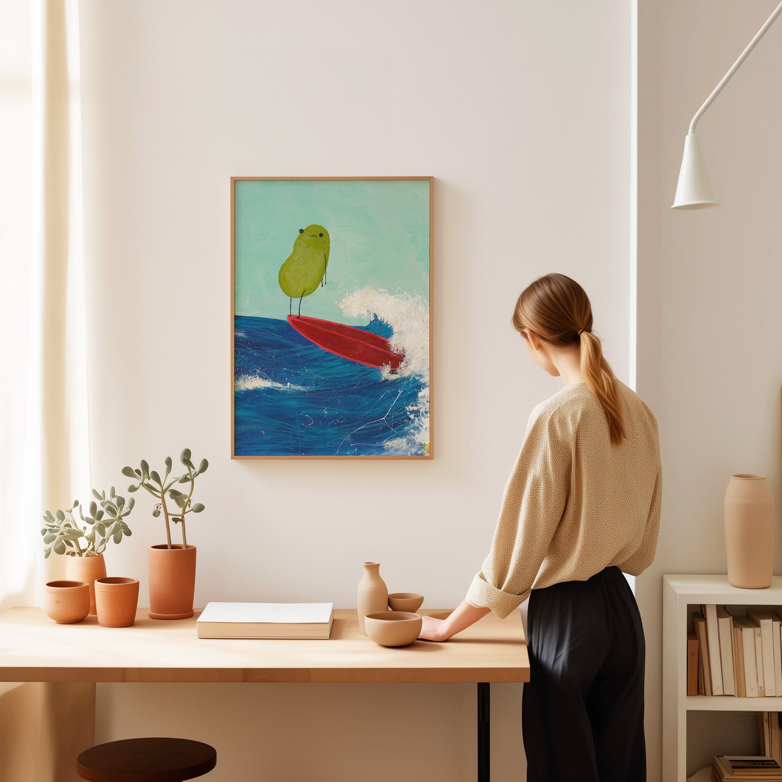 a pickle art print featuring a cute pickle riding a red surfboard on a wave is displayed in a wooden frame over a study desk with a woman viewing the pickle art print