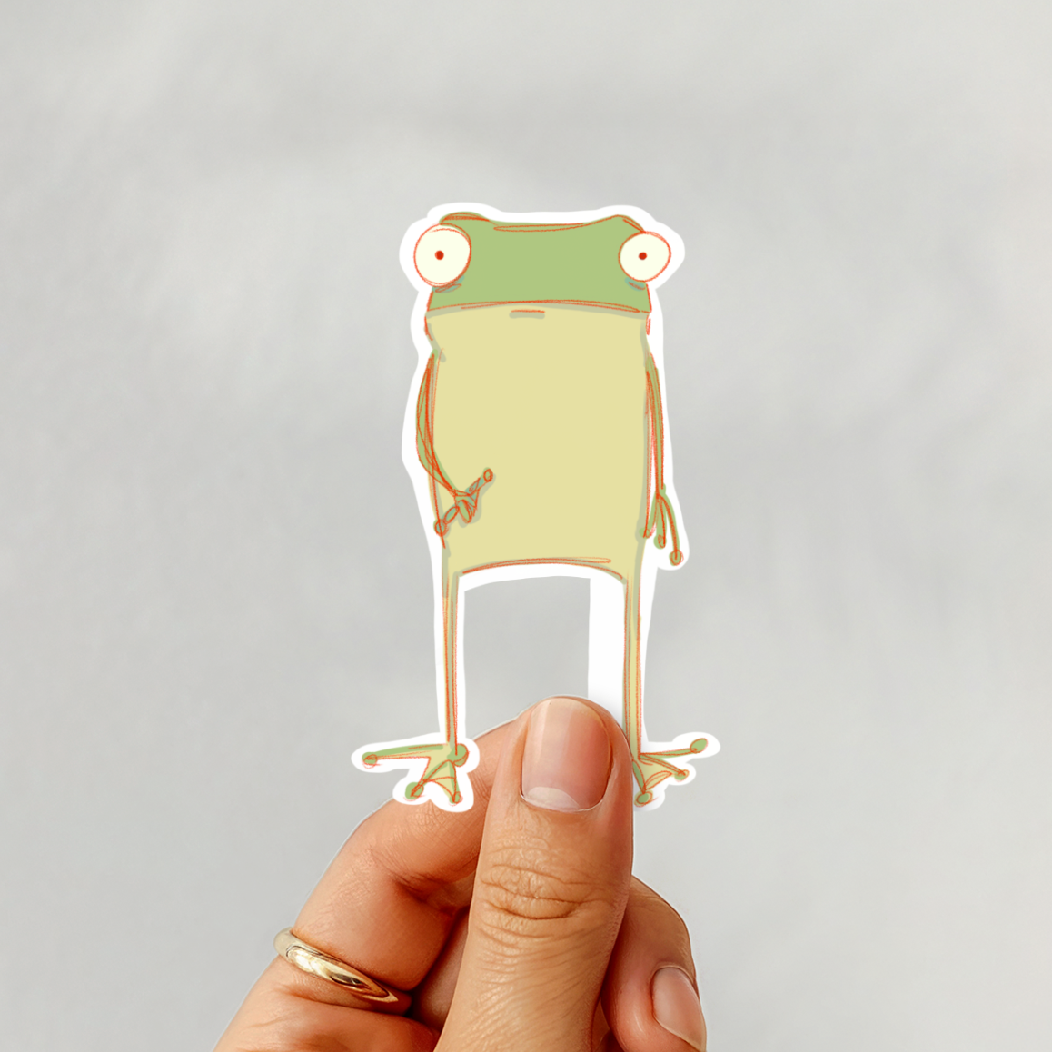 Hand holding a cartoon frog sticker against a plain background