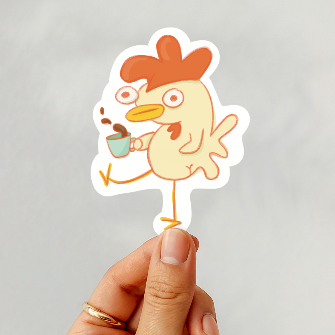 Hand holding a sticker of a cartoon chicken drinking coffee against a gray background