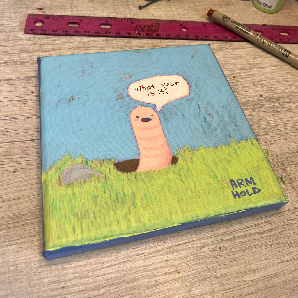 Small painted canvas with a cartoon worm and speech bubble on a wooden surface with art supplies.